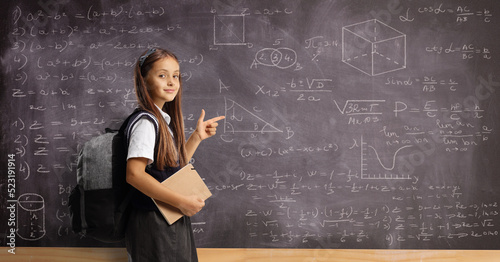 Female pupil in a school uniform with a backpack and book pointing at a blackboard with numbers