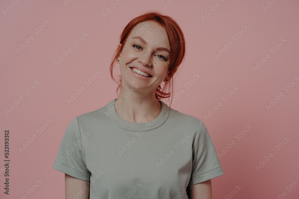 Positive relaxed redhead woman with pleasant appearance smiling cheerfully, isolated on pink