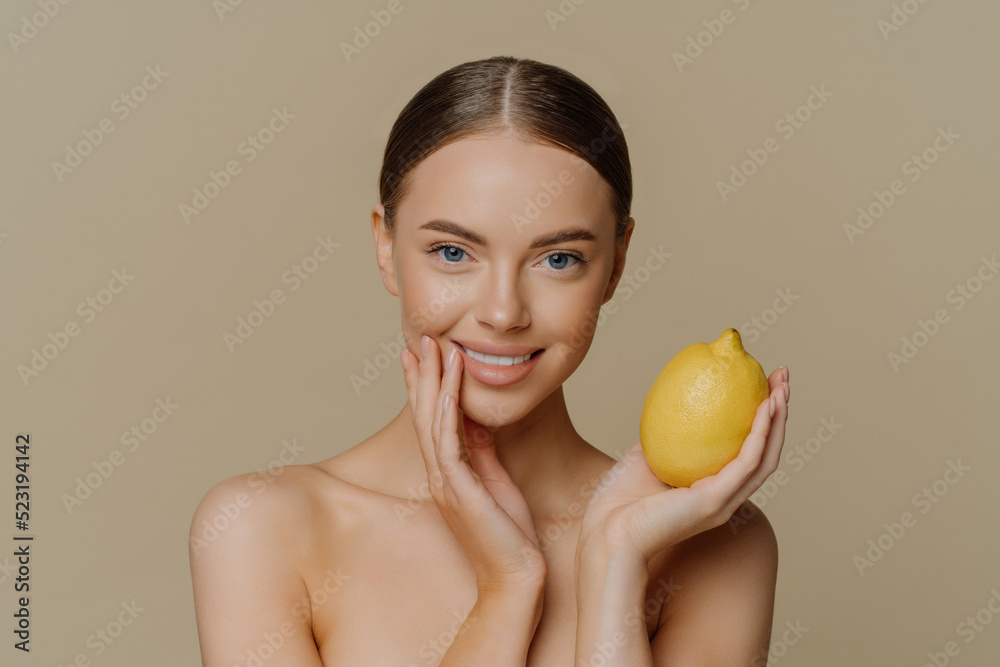 Portrait of charming half naked woman with dark combed hair holds fresh whole lemon uses fruit for natural cosmetics poses against brown background with naked body. People beauty wellness concept Portrait of charming half naked woman with dark combed hair holds fresh whole lemon uses fruit for natural cosmetics poses against brown background with naked body. People beauty wellness concept