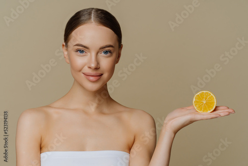 Portrait of lovely dark haired woman holds slice of orange on palm prefers natural beauty products undergoes spa treatment has skin care routine wrapped in bath towel isolated over brown background