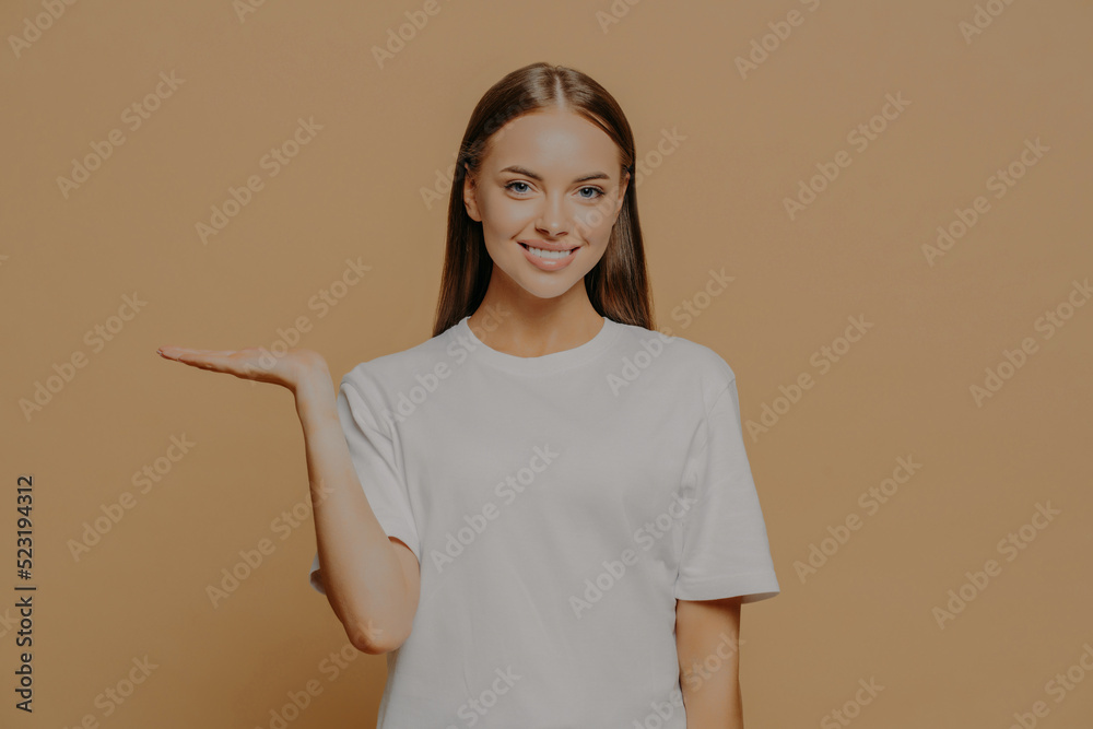 Glad positive young woman raises palm over blank copy space demonstrates invisible object wears casual white t shirt poses against brown background. People advertisement and promotion concept Glad positive young woman raises palm over blank copy space demonstrates invisible object wears casual white t shirt poses against brown background. People advertisement and promotion concept