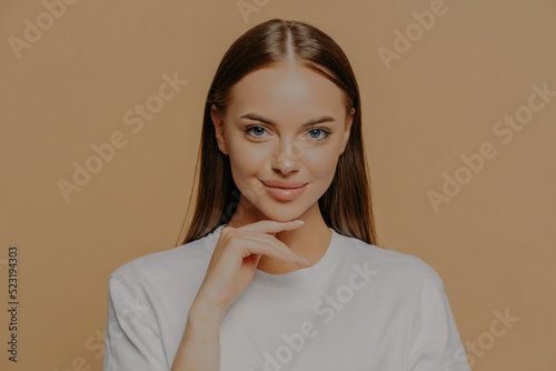 Lovely young European woman with makeup healthy skin touches gently jawline looks directly at camera has long straigh hair natural beauty dressed in casual sweater isolated over brown background photo