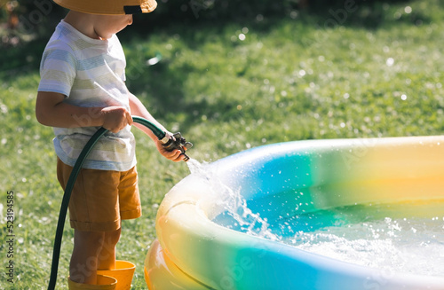 A little boy is playing with a garden hose in the backyard on a sunny day. A preschool child pours an inflatable pool. The concept of raising a child and teaching him to work.