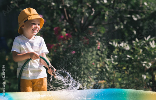 Fototapeta Naklejka Na Ścianę i Meble -  A little boy is playing with a garden hose in the backyard on a sunny day. A preschool child pours an inflatable pool. The concept of raising a child and teaching him to work.