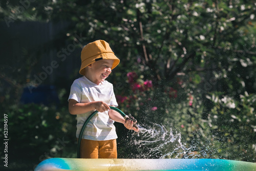 A little boy is playing with a garden hose in the backyard on a sunny day. A preschool child pours an inflatable pool. The concept of raising a child and teaching him to work.