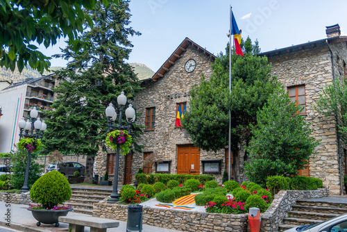 Sant Julia de Loria Town Hall in Andorra, a stone building with the Andorran flag on a mast and a garden with trees and flowers in summer, with the mountains behind and closed wooden doors photo
