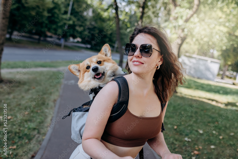 Happy smiling woman traveler is riding her electro scooter in city parkland with dog Welsh Corgi Pembroke in a special backpack Happy smiling woman traveler is riding her electro scooter in city parkland with dog Welsh Corgi Pembroke in a special backpack