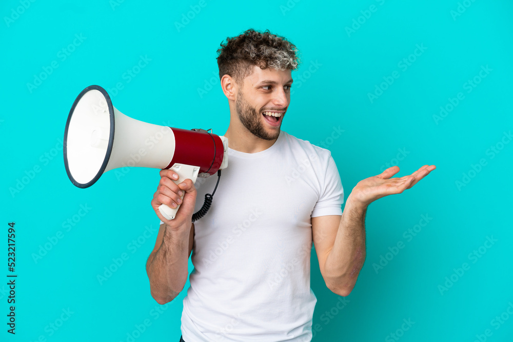 Young handsome caucasian man isolated on blue background holding a megaphone and with surprise facial expression Young handsome caucasian man isolated on blue background holding a megaphone and with surprise facial expression