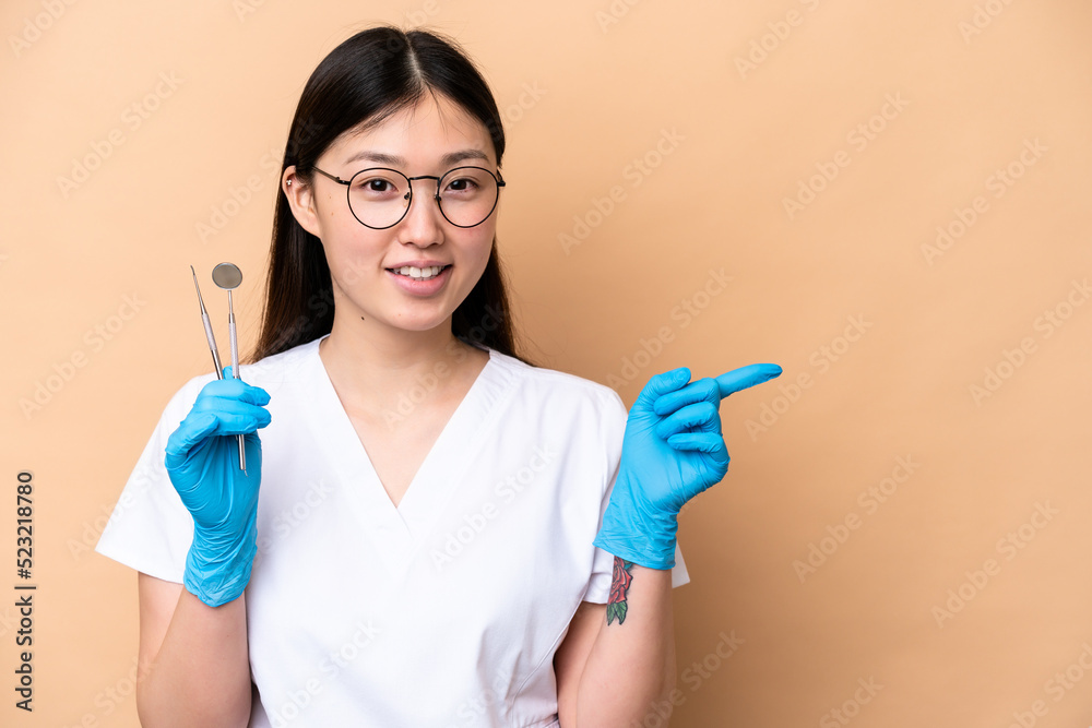 Dentist Chinese woman holding tools isolated on beige background pointing finger to the side Dentist Chinese woman holding tools isolated on beige background pointing finger to the side