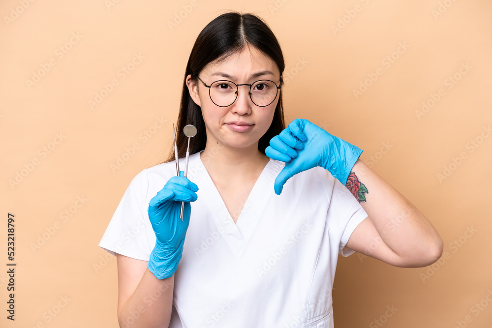 Dentist Chinese woman holding tools isolated on beige background showing thumb down with negative expression Dentist Chinese woman holding tools isolated on beige background showing thumb down with negative expression