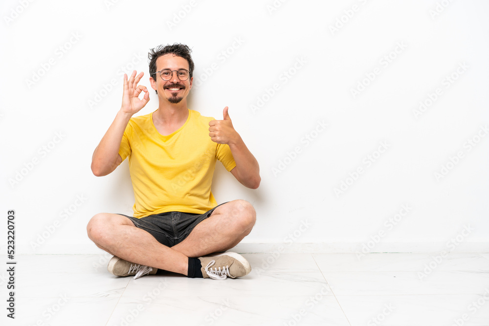Young caucasian man sitting on the floor isolated on white background showing ok sign and thumb up gesture Young caucasian man sitting on the floor isolated on white background showing ok sign and thumb up gesture