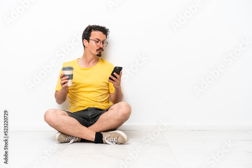 Young caucasian man sitting on the floor isolated on white background holding coffee to take away and a mobile