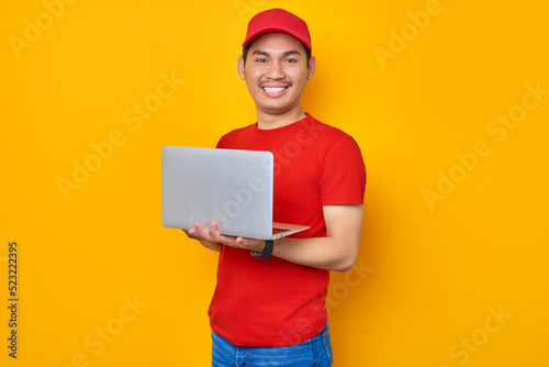 Smiling young Asian man in red cap t-shirt uniform, employee work as dealer courier, using laptop pc computer on work isolated on yellow background. Professional Delivery service concept