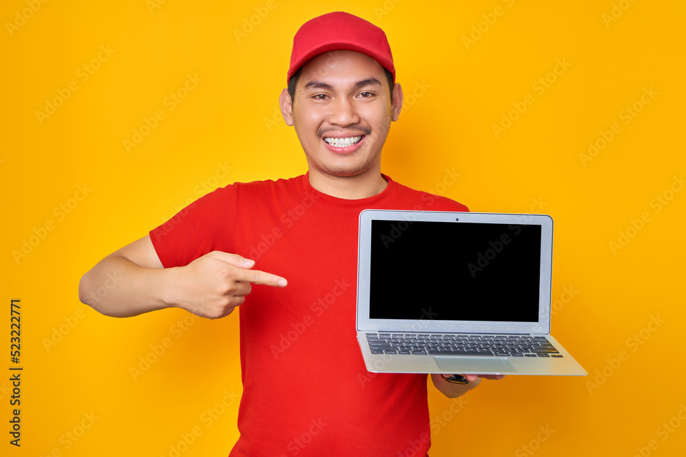 Smiling young Asian man in red cap t-shirt uniform, employee work as dealer courier, pointing fingers at blank screen laptop pc computer on yellow background. Professional Delivery service concept Smiling young Asian man in red cap t-shirt uniform, employee work as dealer courier, pointing fingers at blank screen laptop pc computer on yellow background. Professional Delivery service concept