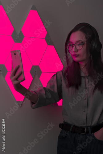 Young pretty woman with wireless headphones in red light of neon panels takes selfie on phone, nightclub and party concept, futuristic neon trend of modern electronic music, dark vertical image