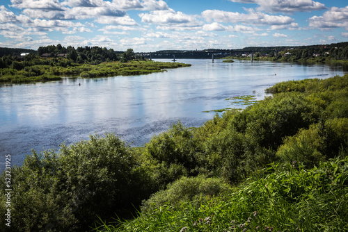 Russia Vologda Oblast Totma 08.12.2017 Summer landscape: coastline the Sukhona River with blue sky and clouds reflecting in calm water, the village, green trees and grass, and the bridge far away