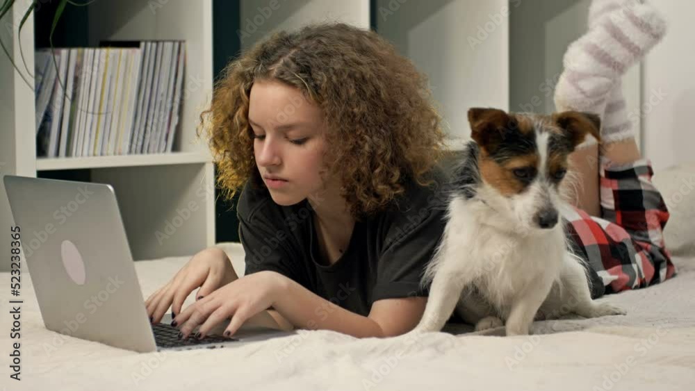 Distance learning. A teenage girl does her homework while lying on the bed with a computer. Beloved dog nearby. Distance learning. A teenage girl does her homework while lying on the bed with a computer. Beloved dog nearby.