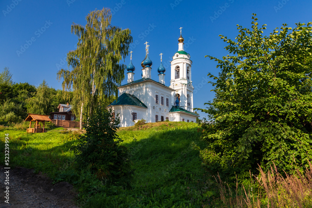 Old Russian Orthodox church under big birch tree on hill top against clear blue sky, Plyos, Russia