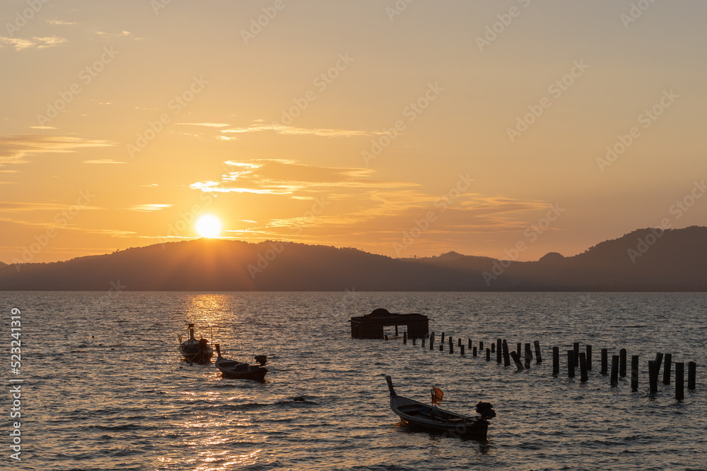 Sunset over tropical island. An old broken pier in the sea with longtail fishing boats moored and the sun setting over the hills of a tropical island