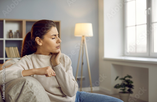 Woman with severe toothache touching her swollen cheek. Young girl feeling discomfort after tooth extraction. Upset lady with inflamed tooth nerve or terrible dental pain sitting on sofa at home photo