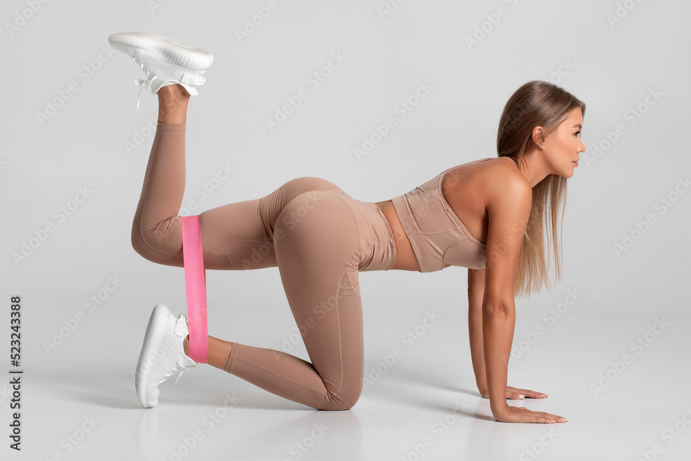 Fitness woman doing kickback exercise for glutes with resistance band on  gray background. Athletic girl working out donkey kicks. Stock Photo