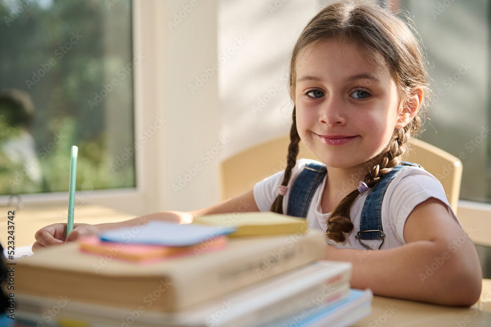 Adorable Caucasian child, schoolgirl cute smiling looking at camera, sitting at a desk with folded textbooks on blurred foreground. Portrait of a happy girl first grader studying in an online school Adorable Caucasian child, schoolgirl cute smiling looking at camera, sitting at a desk with folded textbooks on blurred foreground. Portrait of a happy girl first grader studying in an online school