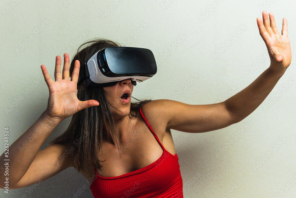 Woman with virtual reality glasses on neutral background raising both hands with amazed face and open mouth. Young girl in her room trying out augmented reality in three dimensions. Woman with virtual reality glasses on neutral background raising both hands with amazed face and open mouth. Young girl in her room trying out augmented reality in three dimensions.