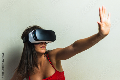 Woman with virtual reality glasses on neutral background raising a hand making the stop gesture. Young girl in her room trying augmented reality in three dimensions.