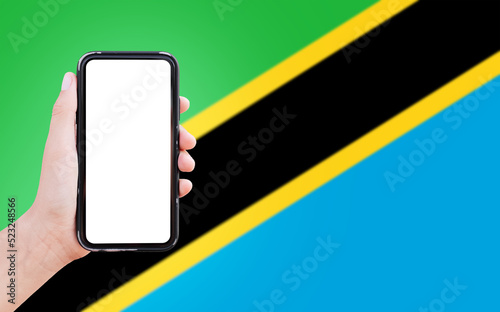 Close-up of male hand holding smartphone with blank on screen, on background of blurred flag of Tanzania.