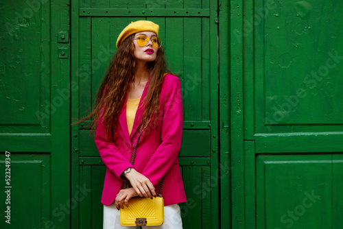 Fashionable confident woman wearing trendy outfit with yellow sunglasses, beret, shoulder bag, pink fuchsia color blazer, posing near green door. Copy, empty space for text