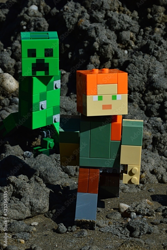 Naklejka premium LEGO Minecraft figure of Alex chased by dangerous green explosive mob creature Creeeper on black sand muddy soil, summer daylight sunshine.