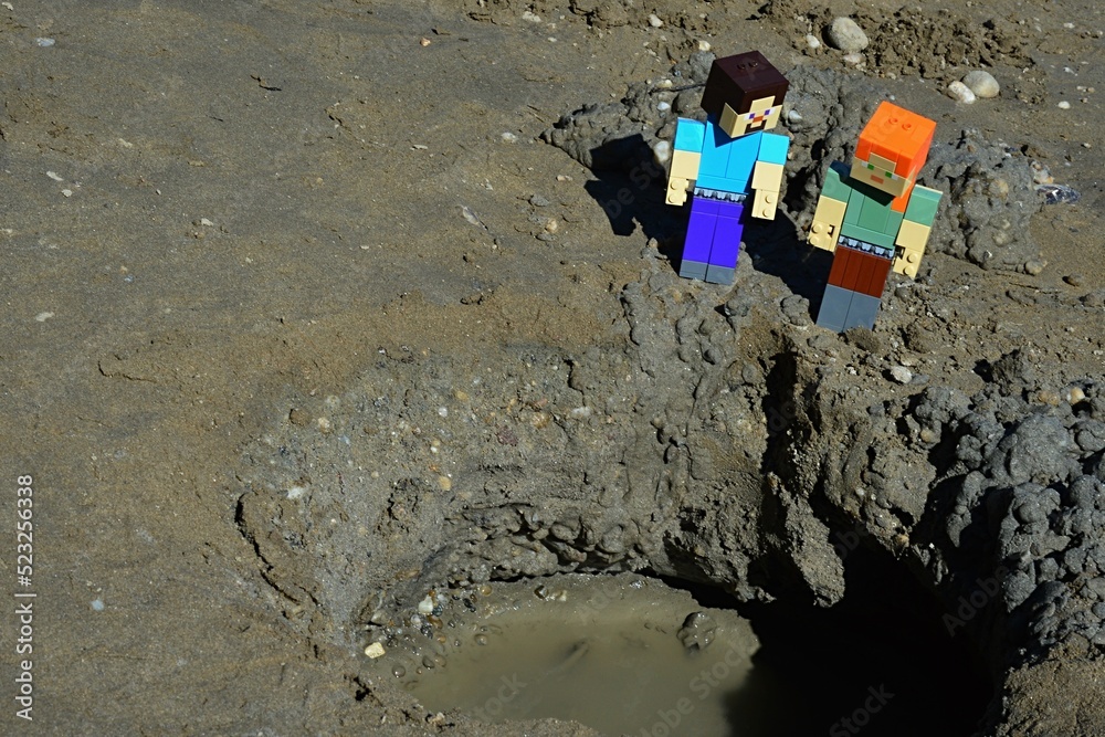 Fototapeta premium LEGO Minecraft large figures of Alex and Steve characters are looking on large wet muddy pit or crater on black sand beach with dirty water on bottom. Summer daylight sunshine.