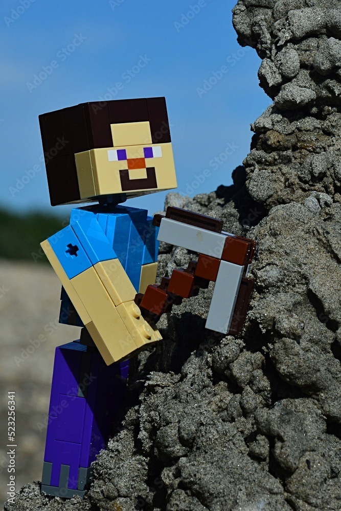 Fototapeta premium LEGO Minecraft large figure of main character Steve with stone pickaxe, climbing on black sand hill to mine some sand, blue summer skies, beach and tree lane in background.