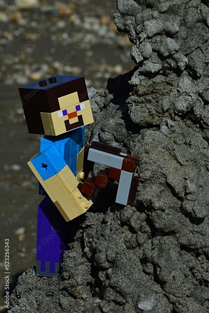 Obraz premium LEGO Minecraft large figure of smiling Steve mining black wet sand from hill on a beach, summer daylight sunshine.