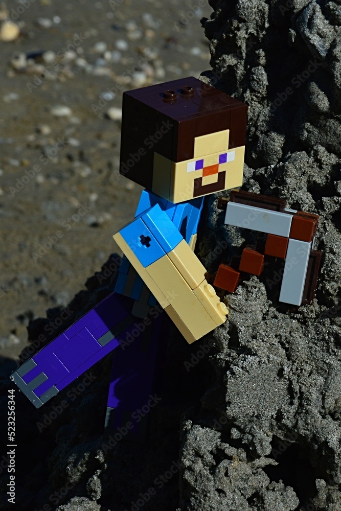 Obraz premium LEGO Minecraft large action figure of Steve with stone pickaxe, ready to mine some wet black sand from steep hill, summer daylight sunshine.