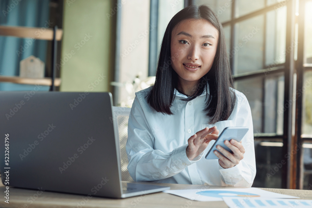 Asian business woman using phone while sitting in office and looking camera Asian business woman using phone while sitting in office and looking camera