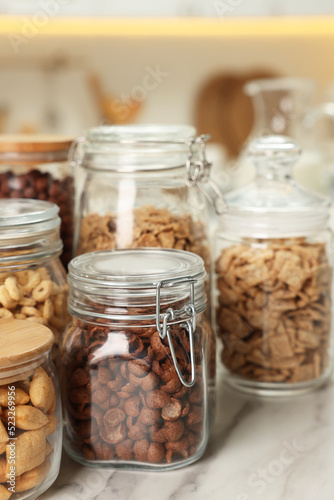 Glass containers with different breakfast cereals on white marble table in kitchen