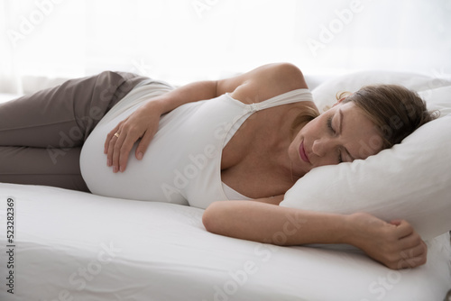 Close up pregnant woman in comfort nightwear sleeps in bed, looking peaceful, touch her big belly enjoy healthy daytime nap, take break in bedroom on fresh white sheets. Late pregnancy, rest concept