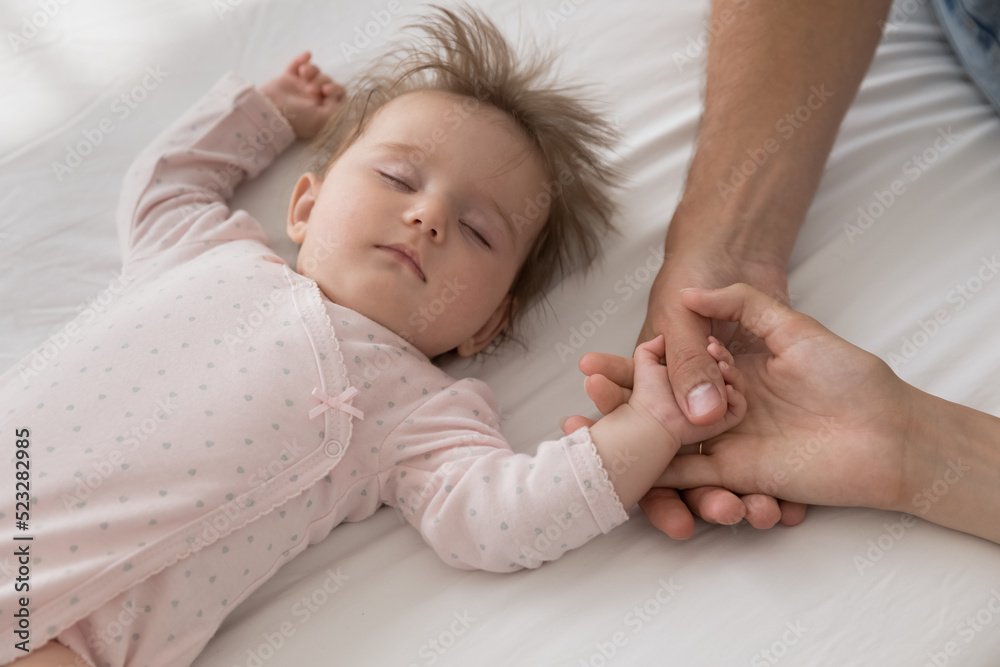Above cropped shot, cute peaceful newborn baby girl in pink bodysuit sleeping on bed holds, touch loving, caring parents hands, close up view. Family bond, ties, unconditional love, adoption concept Above cropped shot, cute peaceful newborn baby girl in pink bodysuit sleeping on bed holds, touch loving, caring parents hands, close up view. Family bond, ties, unconditional love, adoption concept