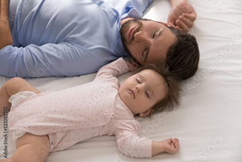 Tired young father falls asleep near newborn baby girl, close up above shot. Sweet infant daughter sleep in bed with loving daddy enjoy daytime nap at home. Fatherhood, family, infancy, rest concept
