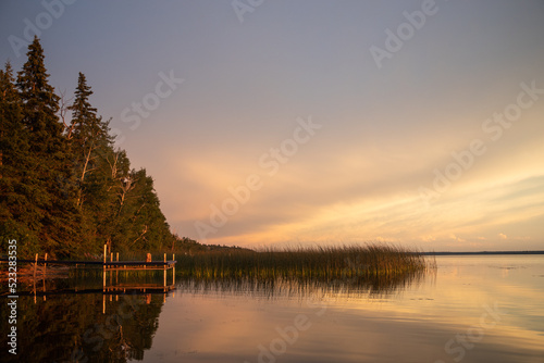 Fototapeta Naklejka Na Ścianę i Meble -  A boat dock by a summer cabin among reeds reflects in the water at sunset in Candle Lake, Northern Saskatchewan, Canada