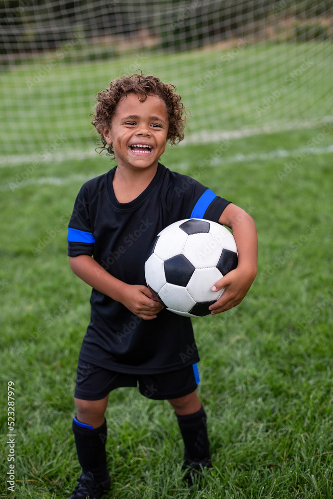 Young African American soccer player standing in front of a soccer goal smiling before a soccer game on a large grass field holding a ball Young African American soccer player standing in front of a soccer goal smiling before a soccer game on a large grass field holding a ball