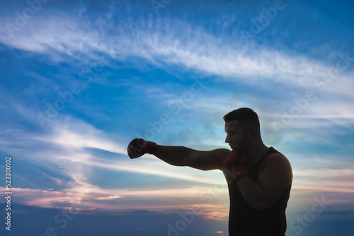 Silhouette of athletic man fighter with boxing gloves posing fight and biceps against blue sky at sunset. Sport, healthy lifestyle and fitness training concept. Copy space