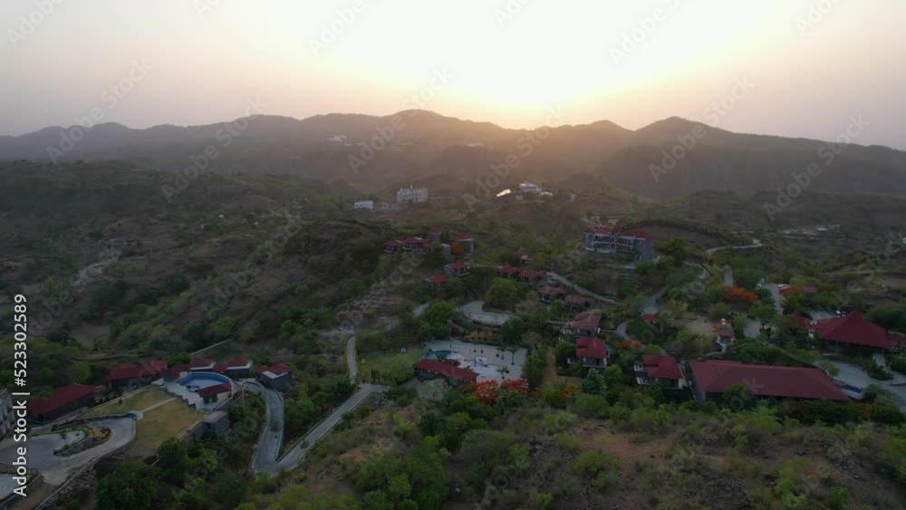 Aerial orbit shot around the sprawling resort complex with red roofs, pool and cottages with the sunset behind the aravali range in the background showing resorts in Kumbhalgarh Rajasthan India