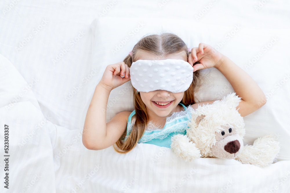 a little girl child with a sleep mask woke up in the morning at home on a bed on a white cotton bed with a teddy bear toy and a cute smile a little girl child with a sleep mask woke up in the morning at home on a bed on a white cotton bed with a teddy bear toy and a cute smile