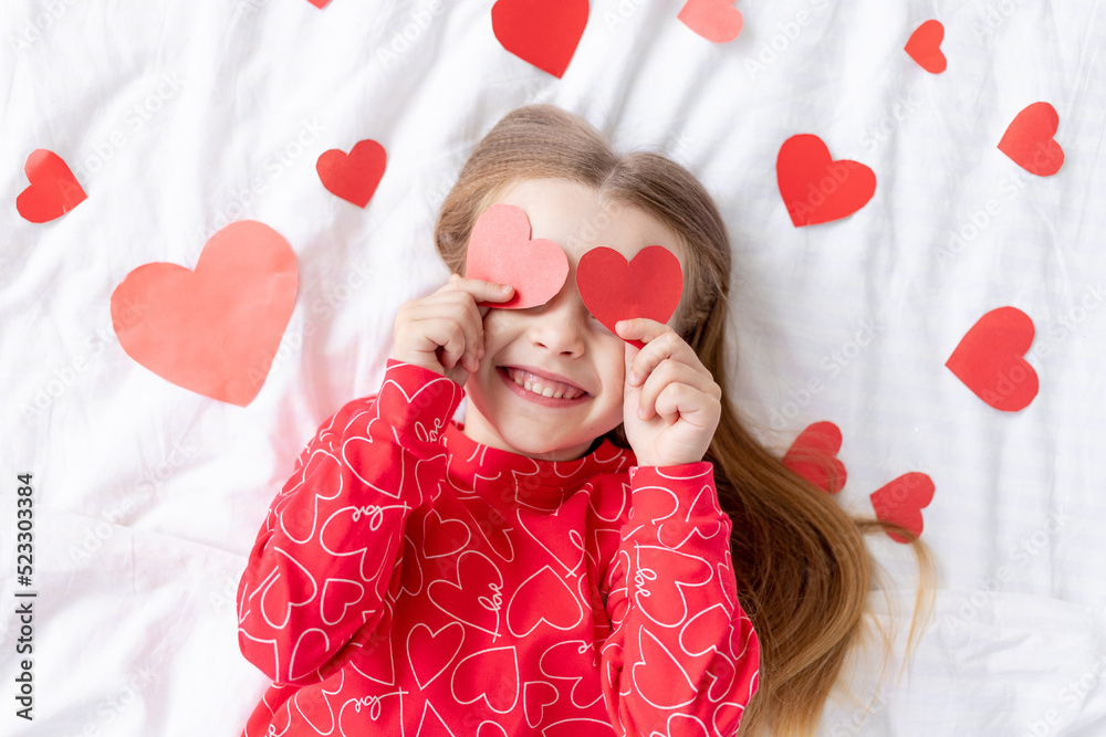 the concept of Valentine's day, a sweet child girl lying on a white bed at home in red pajamas among hearts covers her face with hearts, indulges and fools around, smiles with happiness the concept of Valentine's day, a sweet child girl lying on a white bed at home in red pajamas among hearts covers her face with hearts, indulges and fools around, smiles with happiness