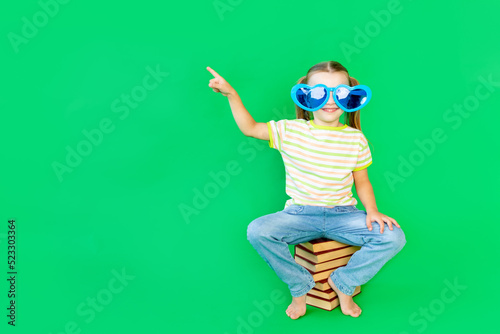 a girl child sits on a green background on books in blue large funny glasses and points her finger up or to the side, space for text