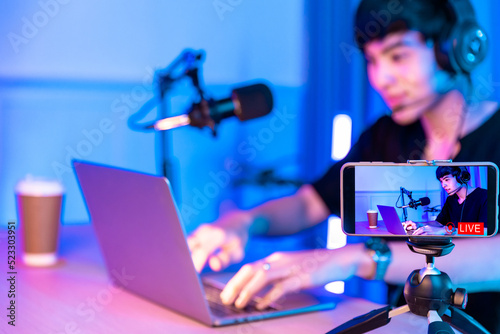 Playing video games on smartphone. Young asian handsome man sitting on chair holding cellphone in his hand. Exited streamer wearing headset in online mobile game in neon room .