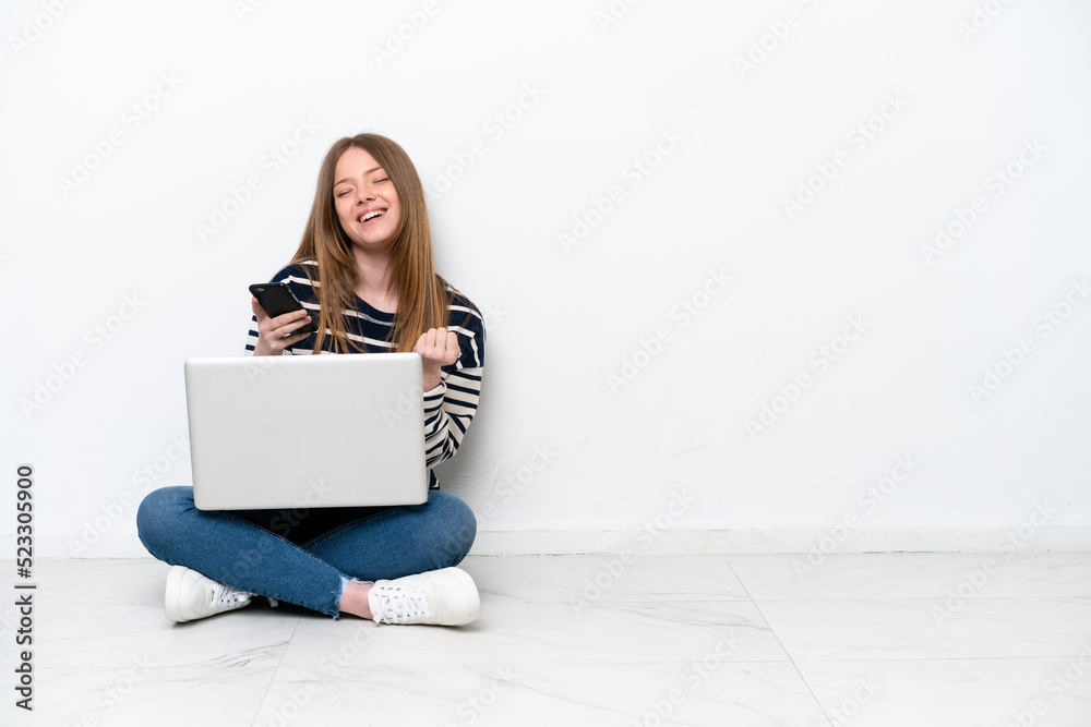 Young caucasian woman with a laptop sitting on the floor isolated on white background with phone in victory position Young caucasian woman with a laptop sitting on the floor isolated on white background with phone in victory position