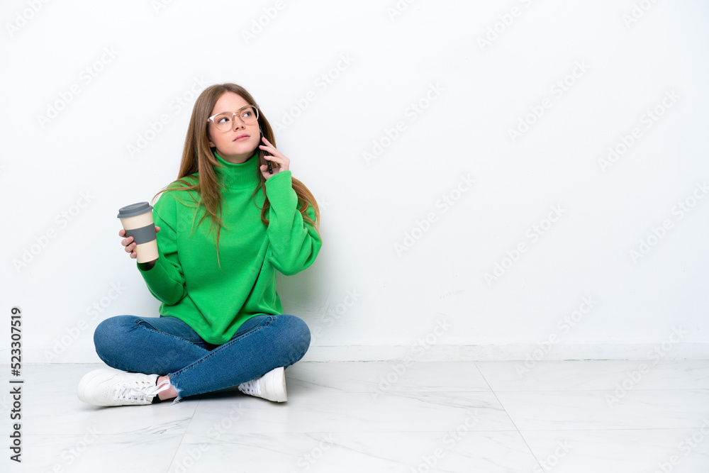 Young caucasian woman sitting on the floor isolated on white background holding coffee to take away and a mobile Young caucasian woman sitting on the floor isolated on white background holding coffee to take away and a mobile